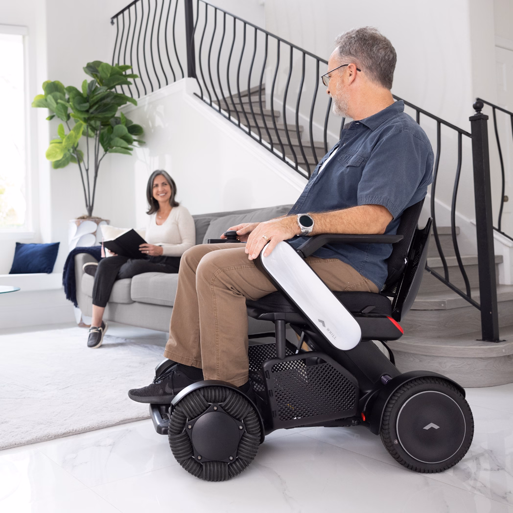 Man using a motorized wheelchair indoors, woman sitting in the background.